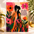 A hardcover journal featuring a colorful floral illustration and a black silhouette of a woman, with the text 'SHE GROWS IN SILENCE. SHE BLOOMS ON PURPOSE.